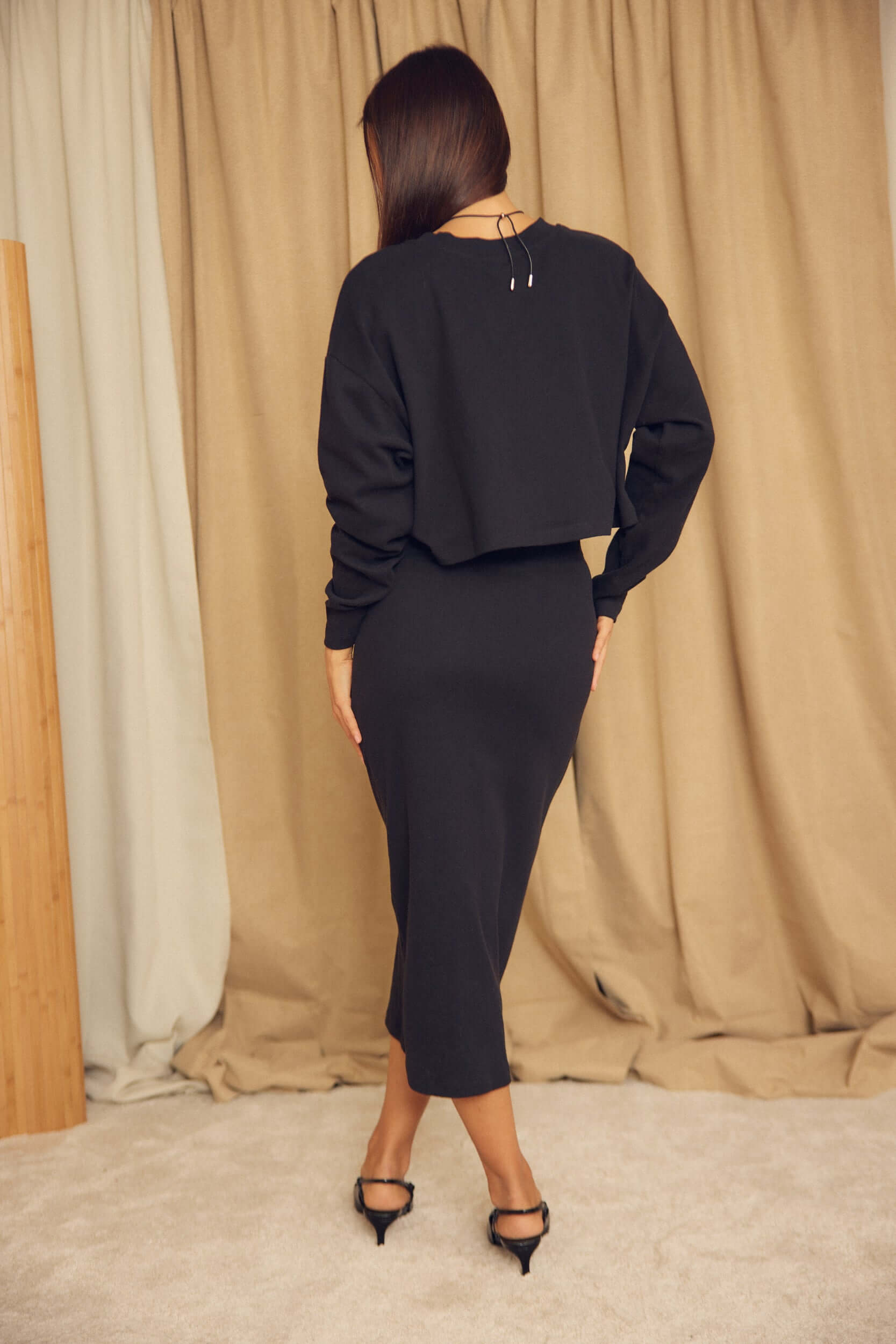 Black Organic Cotton Pencil Midi Skirt paired with a cropped black sweater, modeled from behind on a soft carpet.